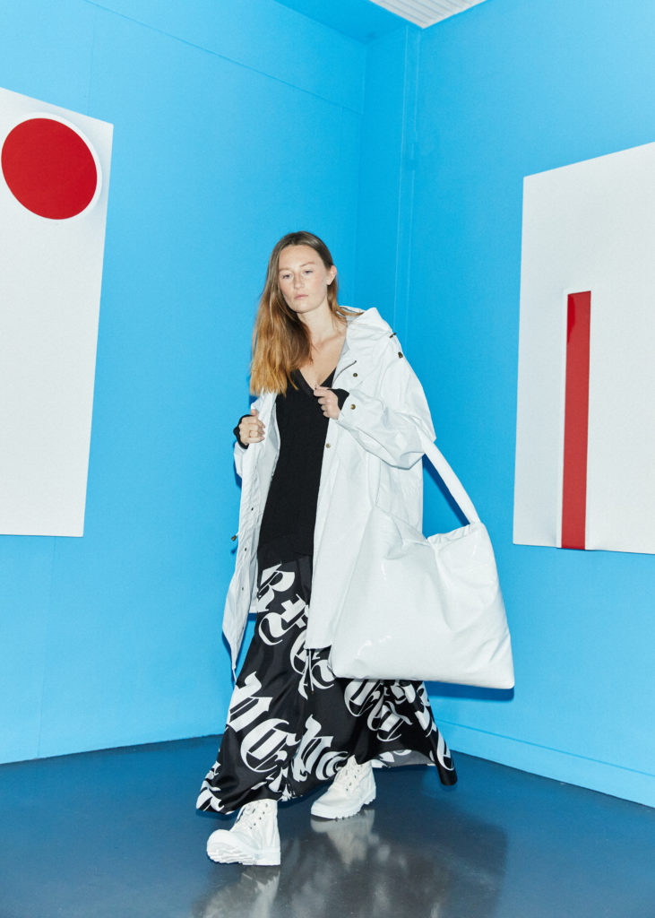 Model wears long black v-neck top undernear a white mack and oversized white handbag, along with white sneakers and long black skirt emblazoned with 'Perfect' written in a gothic script, all by Nomad Atelier