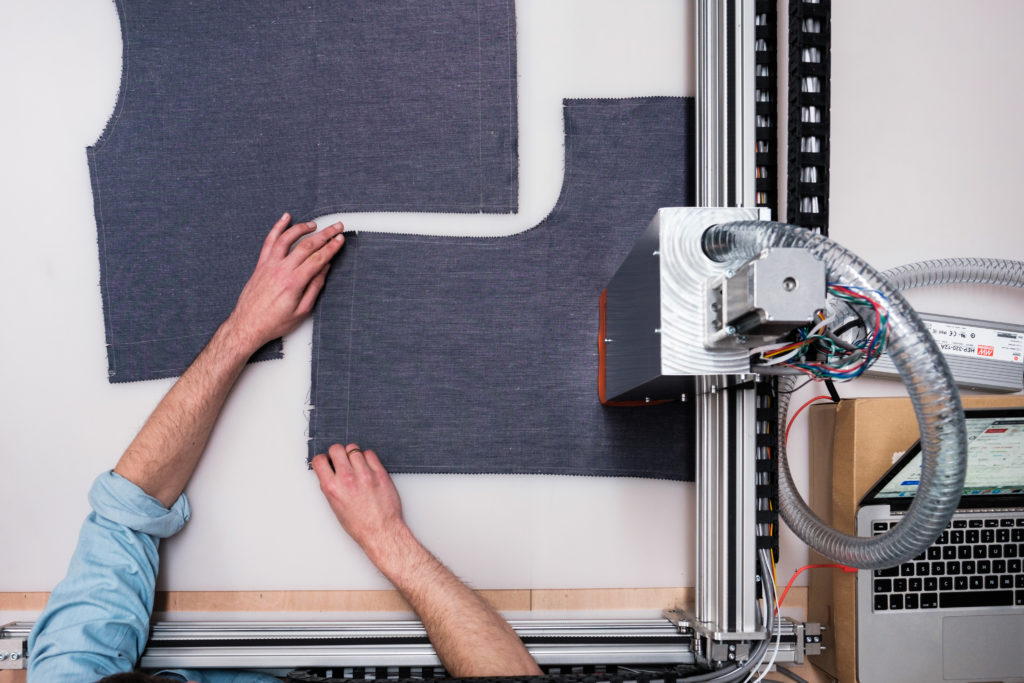 Panel of navy blue fabric seen from above being cut by a silver metallic box on a rail (the cutting machine)