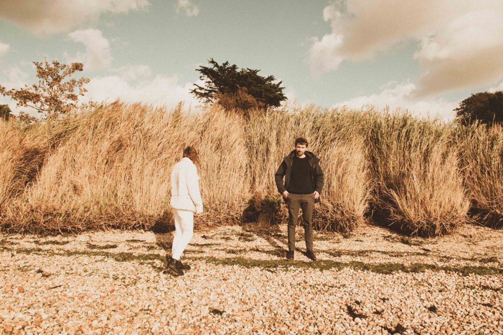 A woman with her back to the camera and a man facing the camera, wearing insulated jackets against a backdrop of high reeds and grass