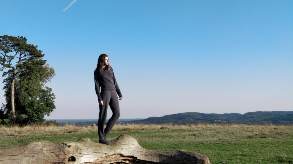 Woman wearing dark grey long-sleeve top, trousers and socks stands on a fallen tree in the middle of open countryside against a wide blue sky