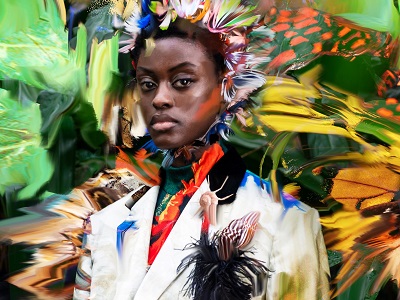 A model wears a high-necked top and tailored jacket in a variety of bright colours against a brightly coloured background of natural greens, yellows, and oranges